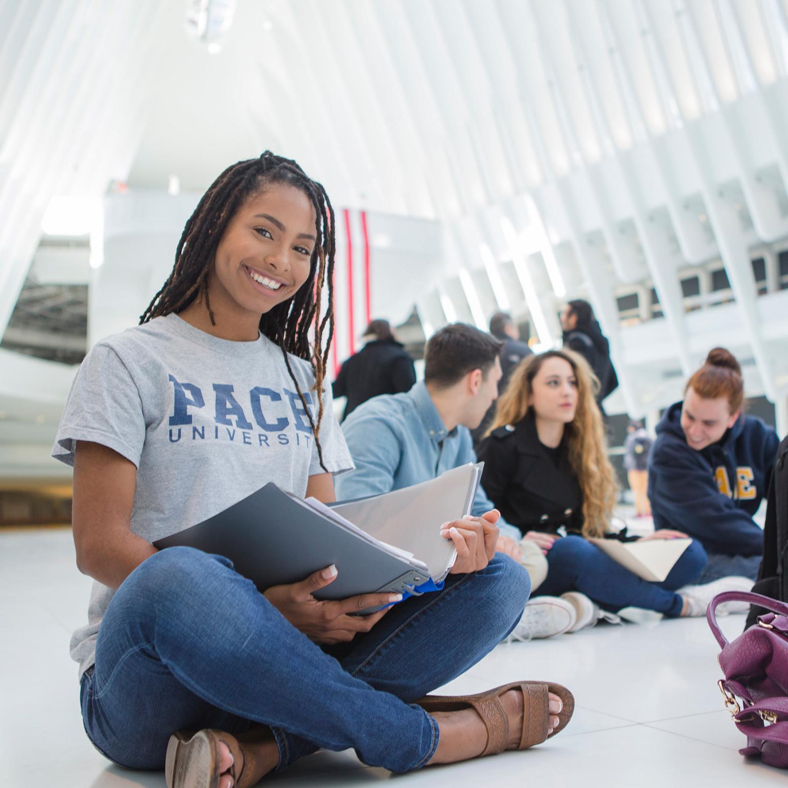 Student sitting on the floor holding a book and smiling at the camera.