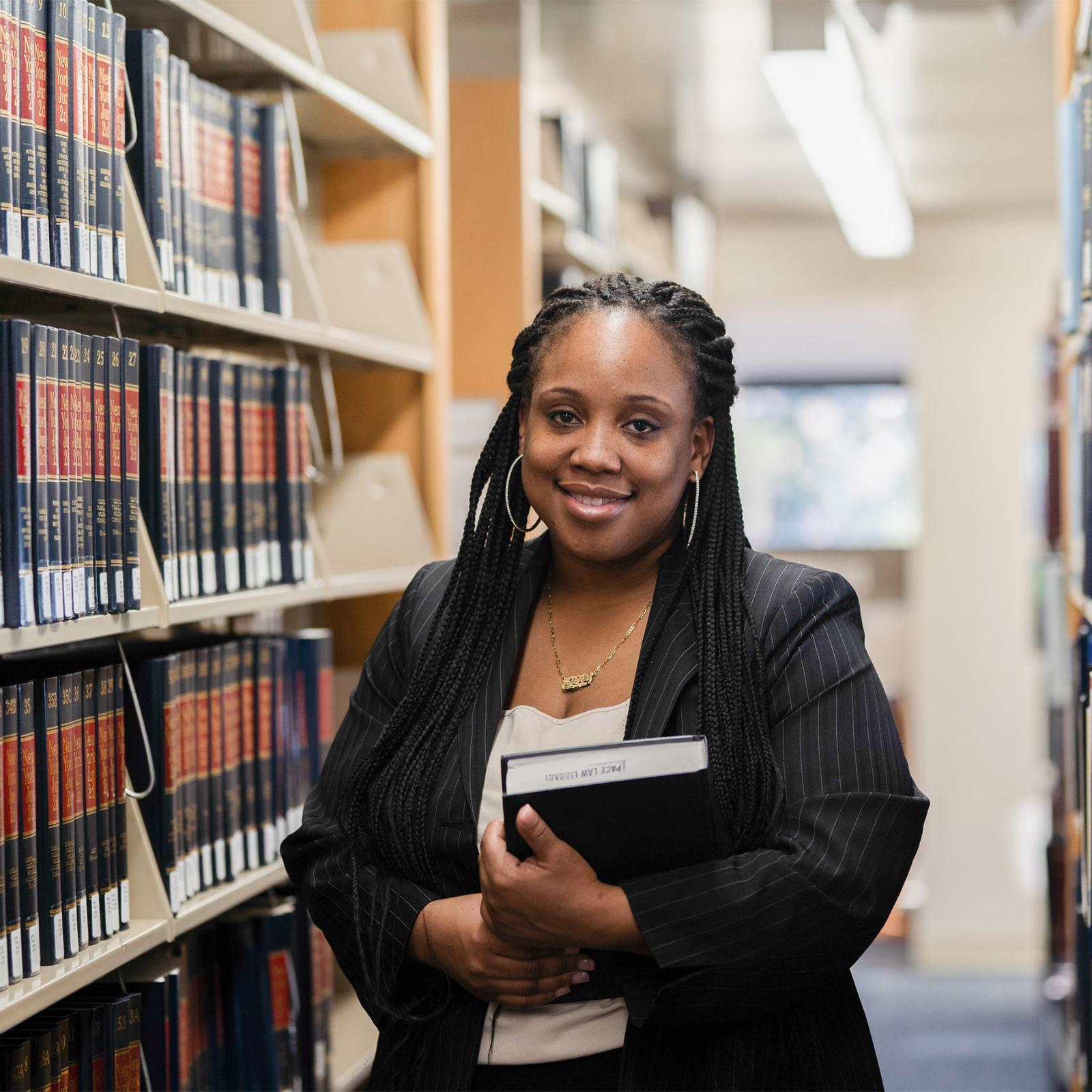 Female student standing in the law library, holding a book and smiling at the camera.