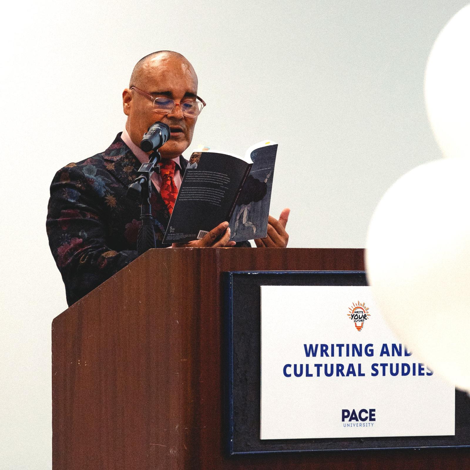 A male reading a book at a podium at an event for the Pace University Writing and Cultural Studies department