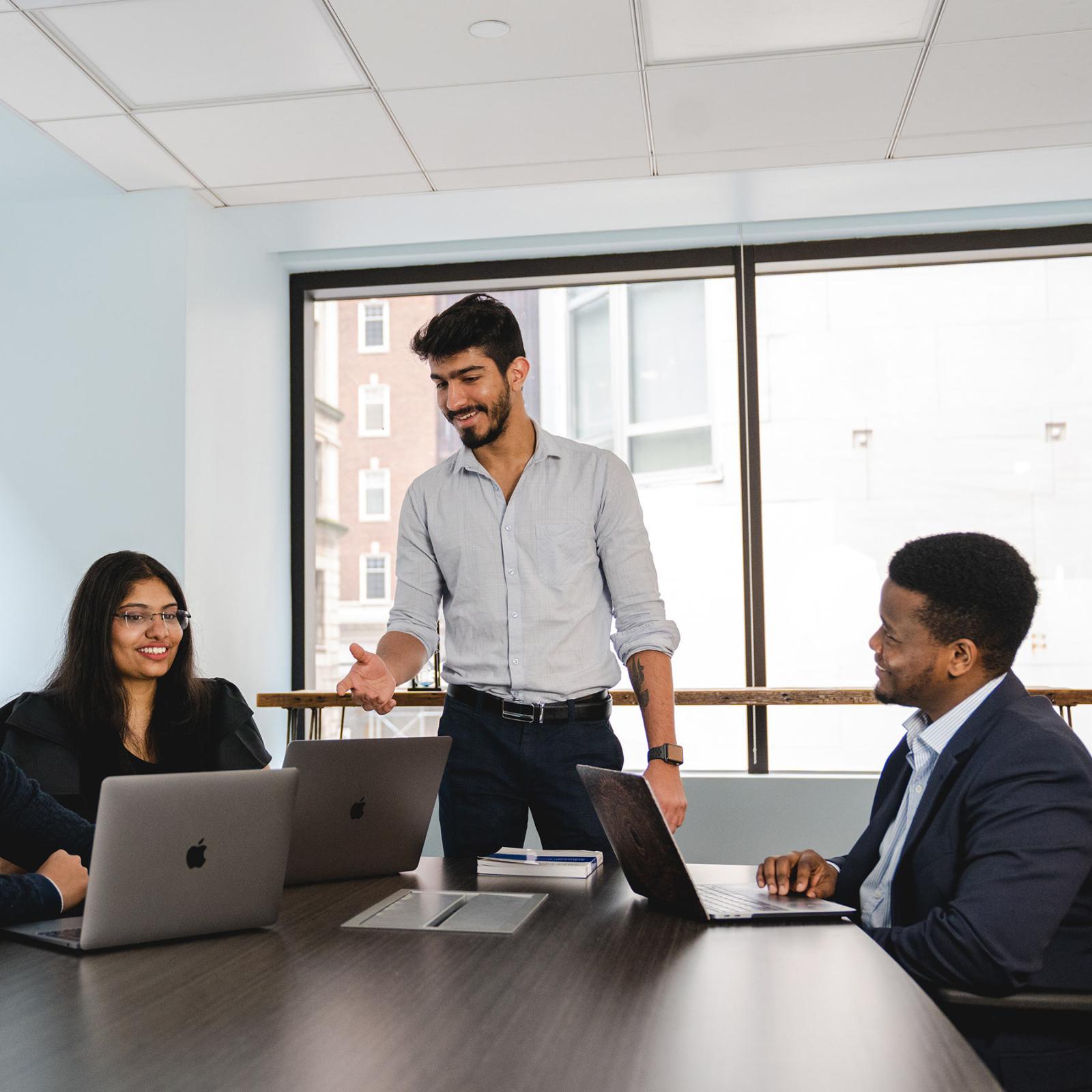 Group of people in business attire sitting at a desk with their laptops smiling.