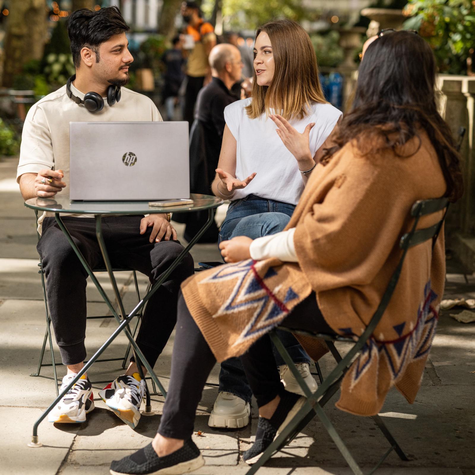 three people sitting at an outdoor table with an open laptop