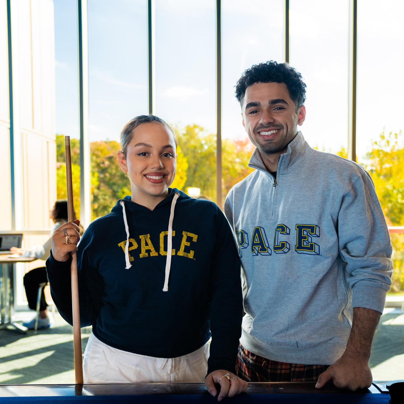Two Pace students stand in front of a pool table holding one of the sticks