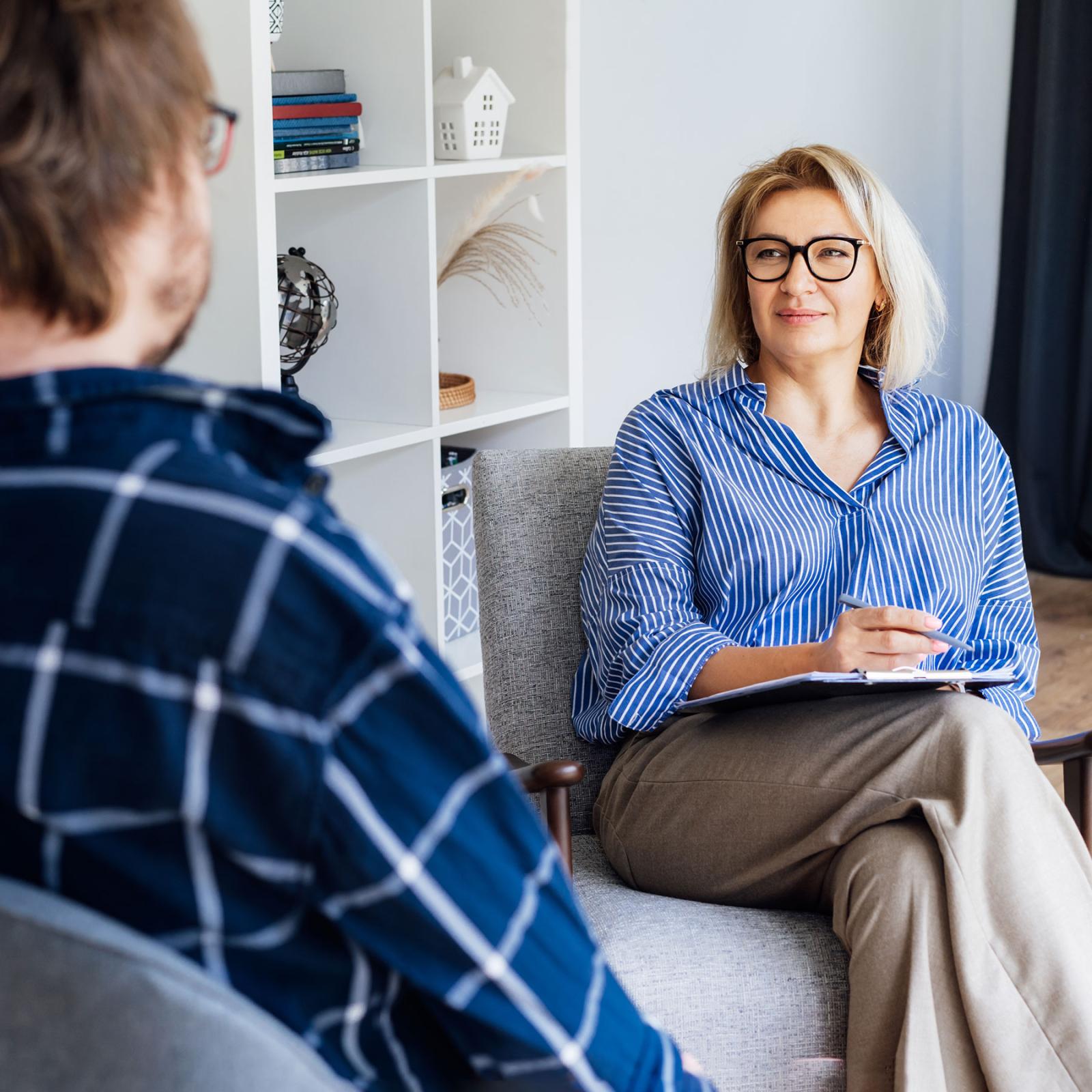 Female with a clipboard facing a male in a therapy session