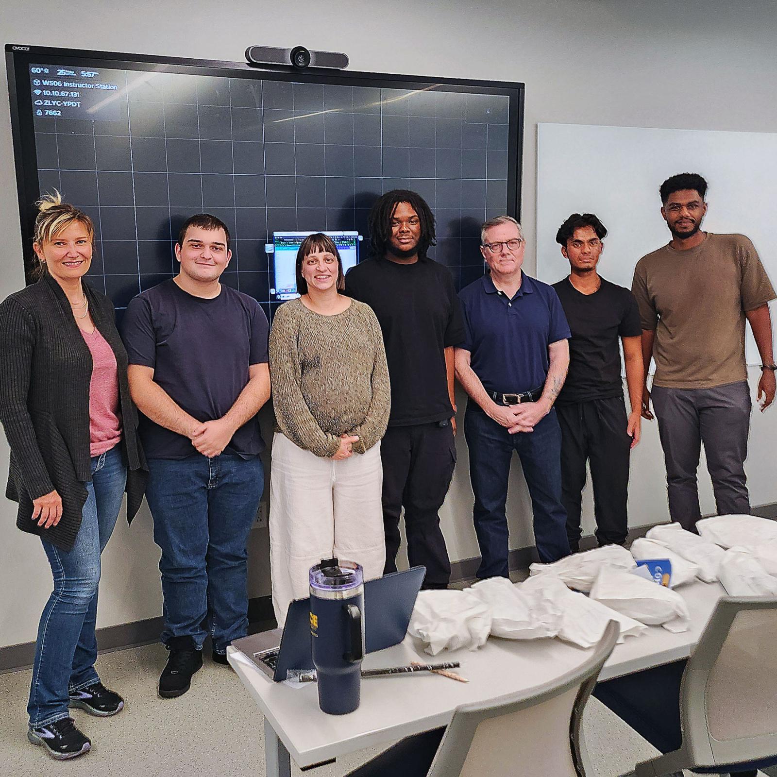 The Economics Hackathon winning group standing with faculty in front of a screen with a table and chairs in front of them