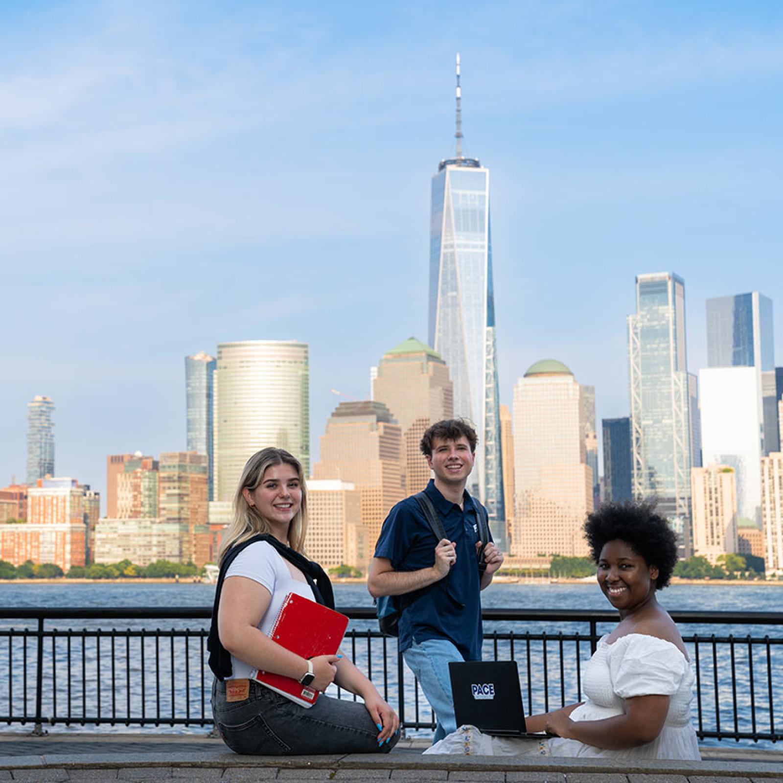 Three Pace students socialize near the waterfront, the NYC skyline behind them