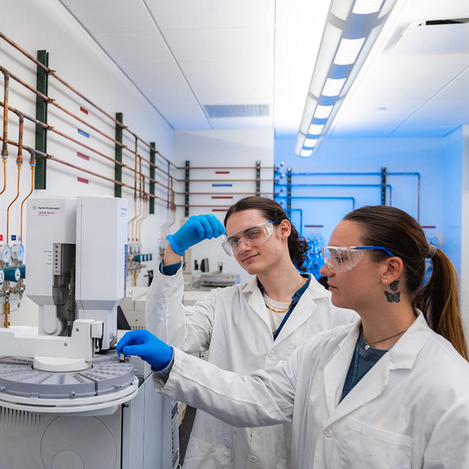 Two Pace University students in lab coats observe lab equipment.