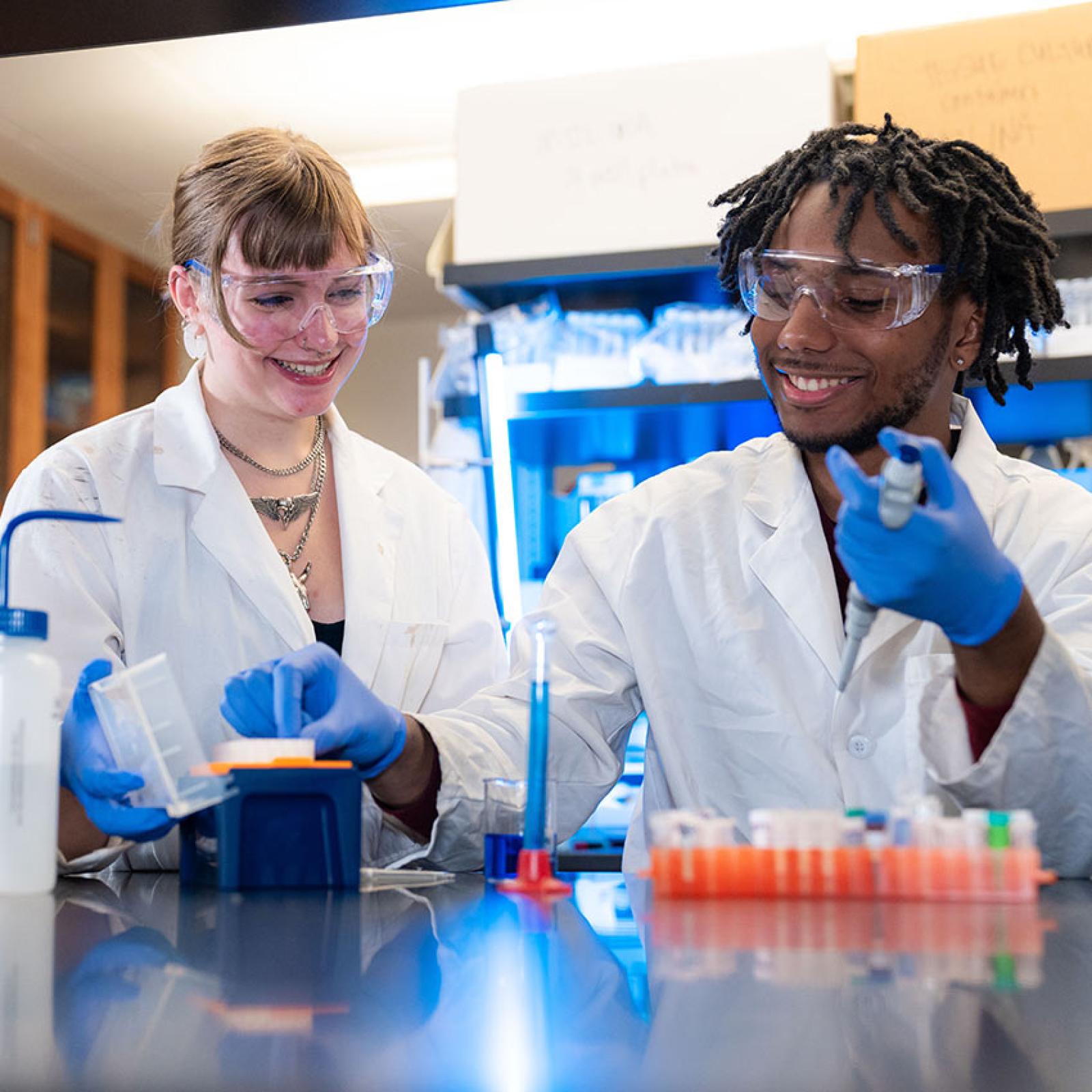 Two Pace University students wear lab coats and protective gear as they do lab research.