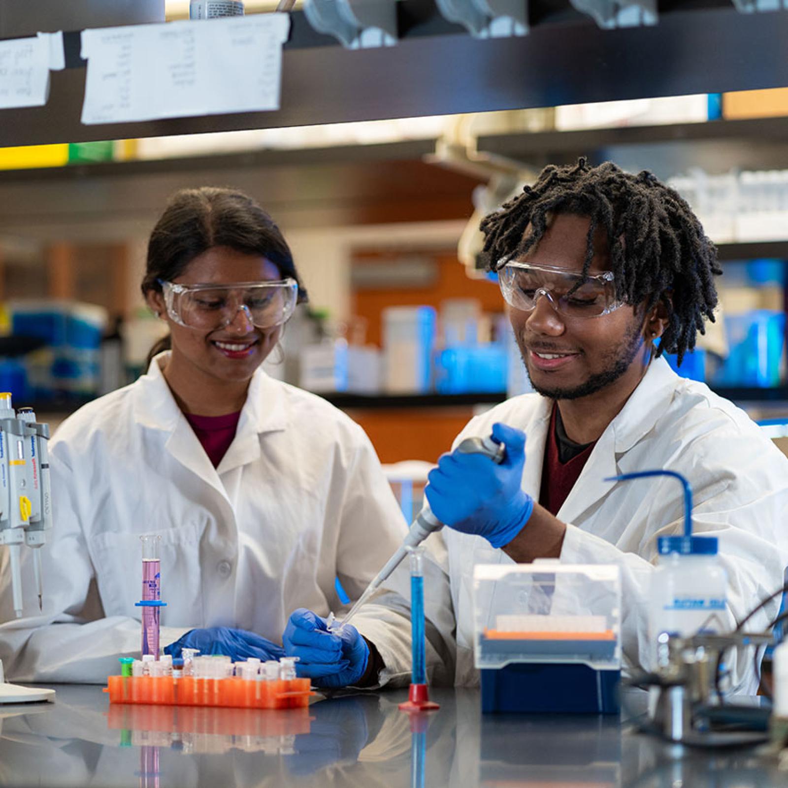 Two Pace University students wear lab coats and goggles during an experiment in the lab.