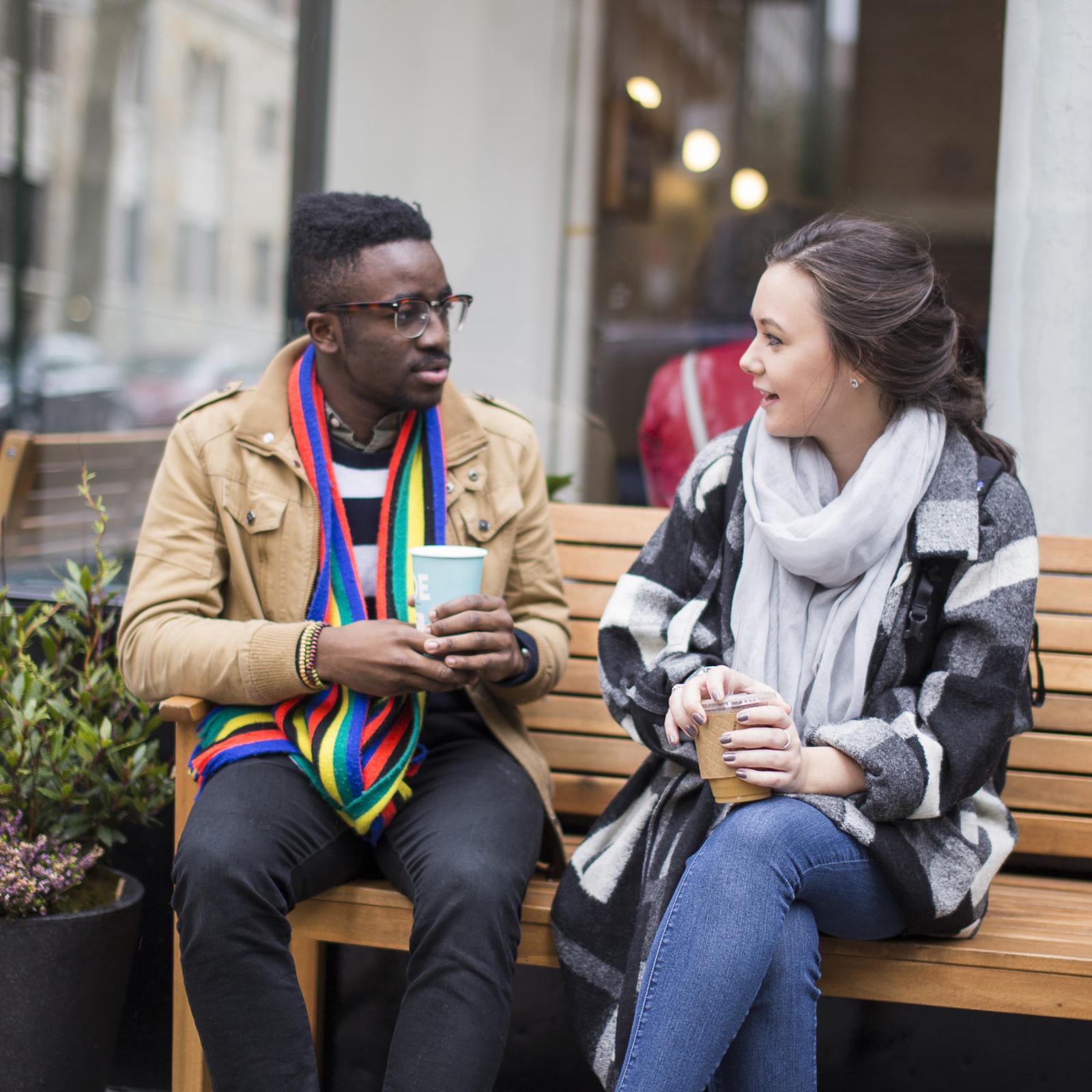 Two Pace students sitting on a bench with coffee discussing psychology.
