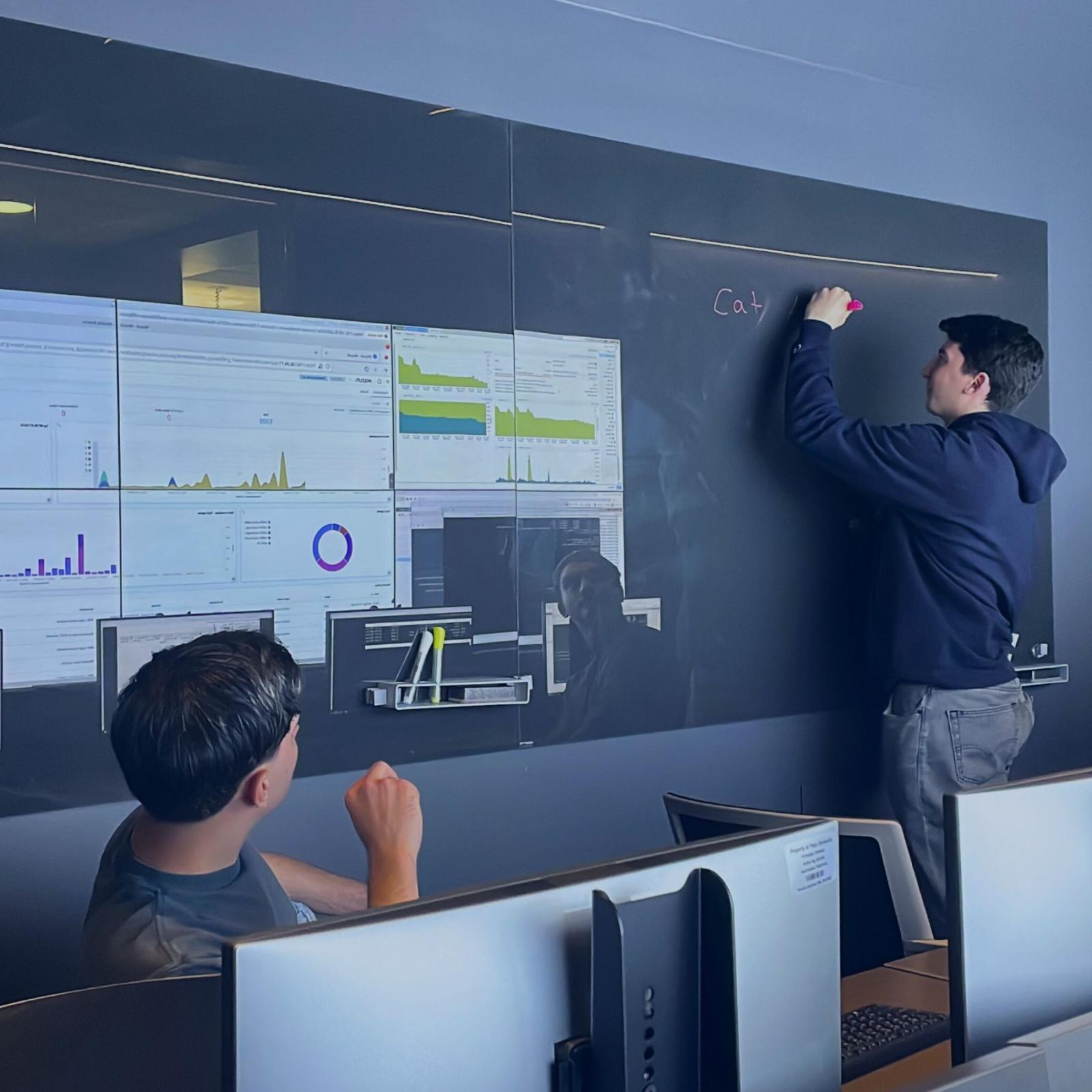 Two Seidenberg students working in the Pace Cyber Range. One of them is writing information on a blackboard, while the other is observing. The screen of the cyber range is visible in the background.