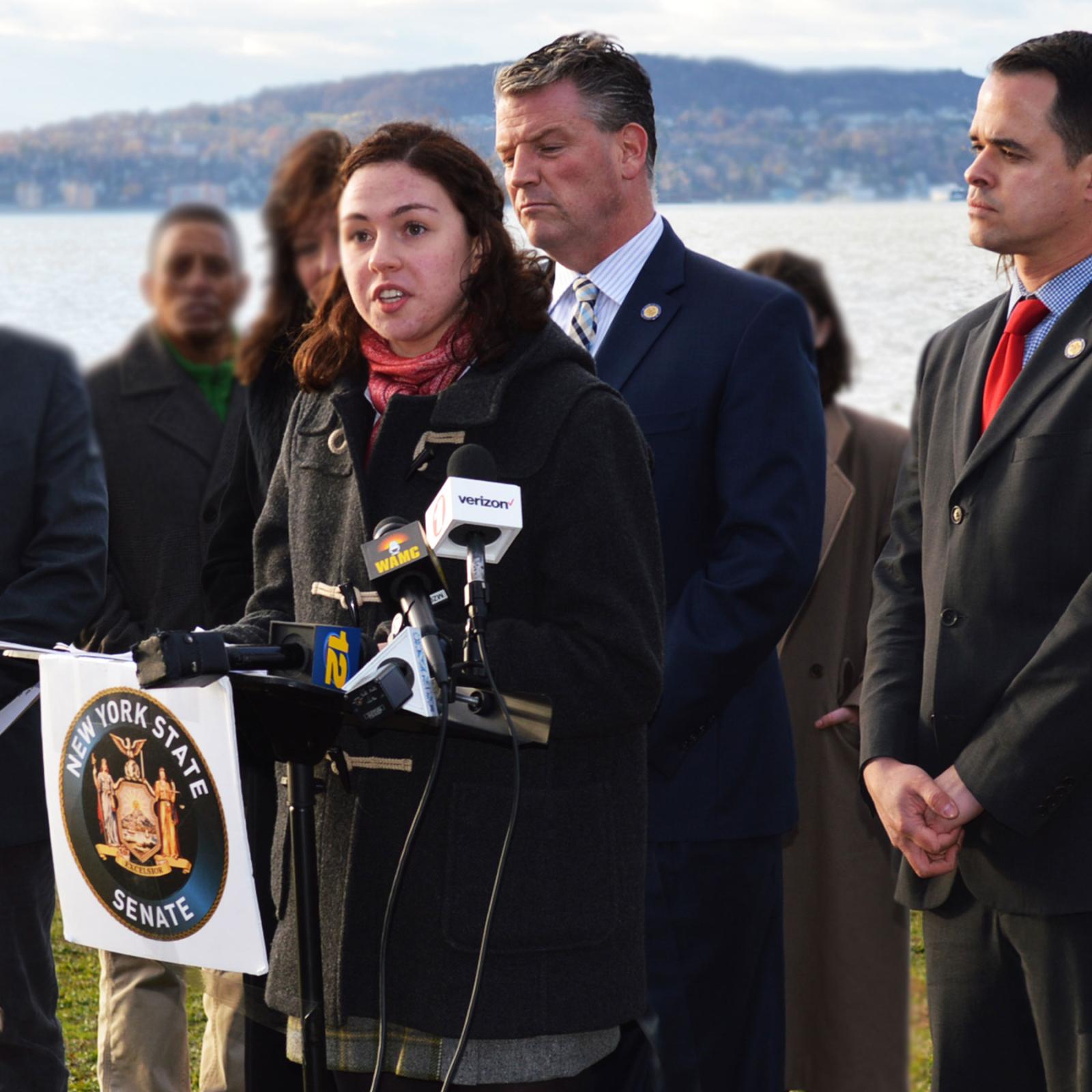 Female speaking with media at a podium with New York State Senate seal on front