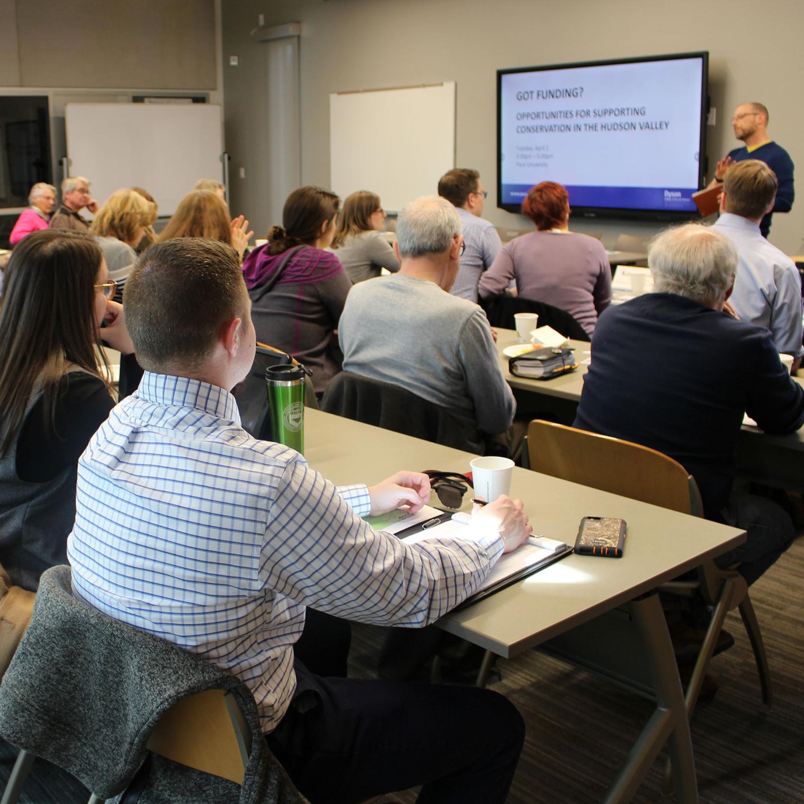 Group of professionals sitting at 3 rows of tables listening to a Pace University program lecture and looking at a powerpoint