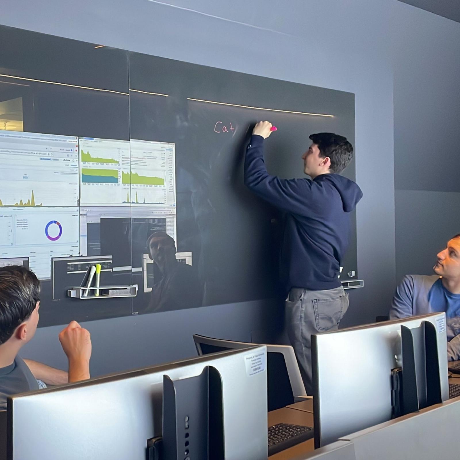 Three Seidenberg students working in the Pace Cyber Range. One of them is writing information on a blackboard, while the others are observing. The screen of the cyber range is visible in the background.