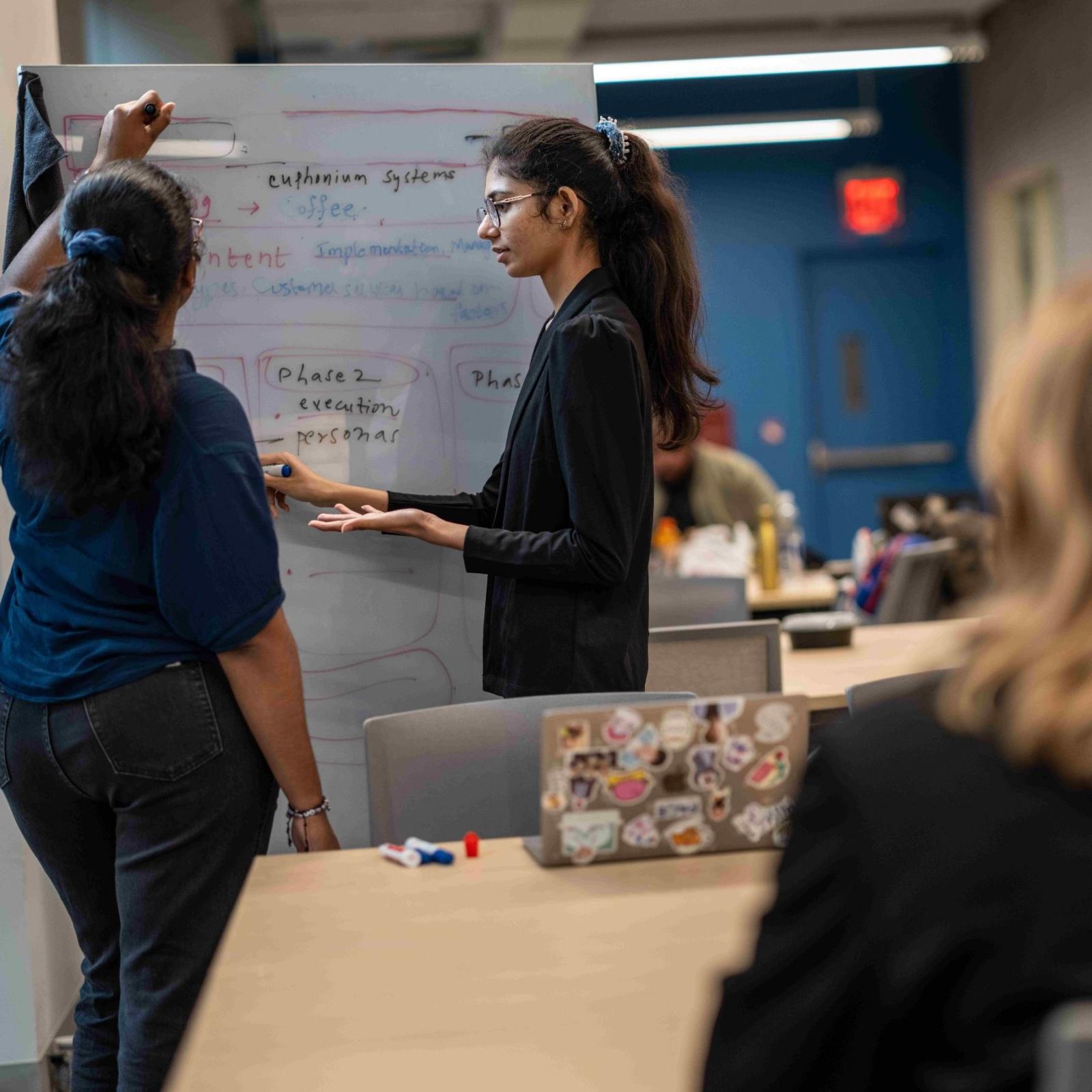 Two Seidenberg students writing on a whiteboard while a professor watches them work.