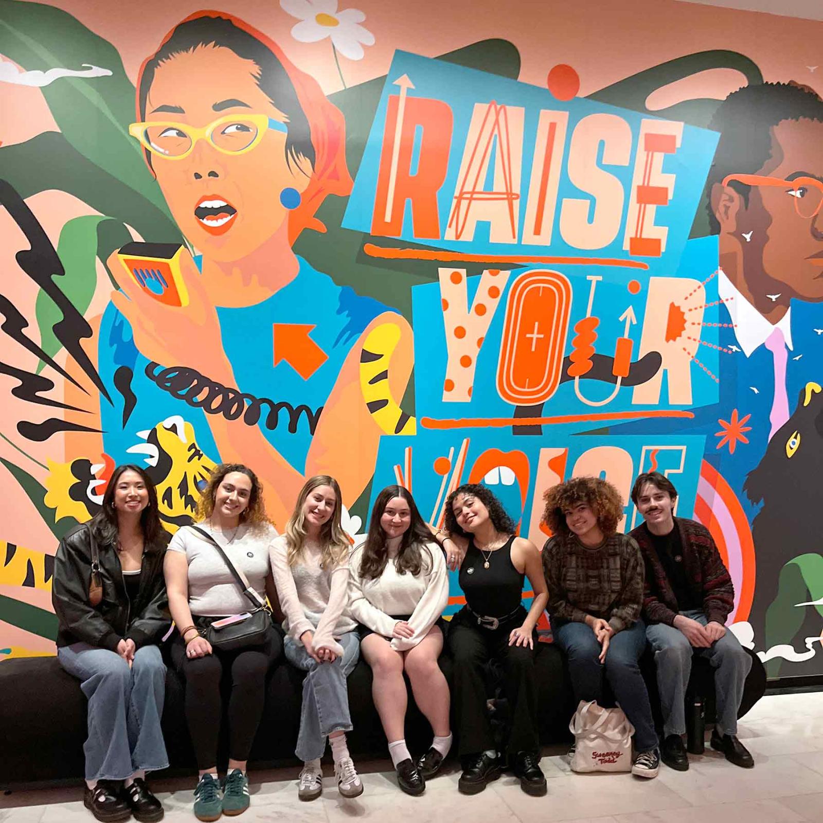Group of Pace University students sitting in front of a Raise Your Voice mural