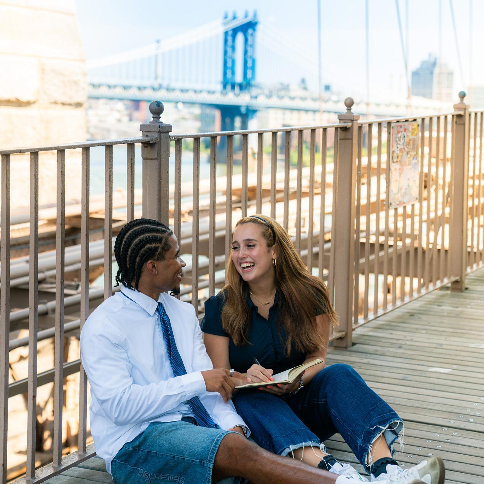 Two Pace University students sitting on the ground in NYC, in front of the Brooklyn Bridge.