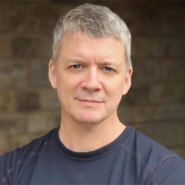 Portrait of Chris Coffey wearing a dark blue shirt and a tan bricked background