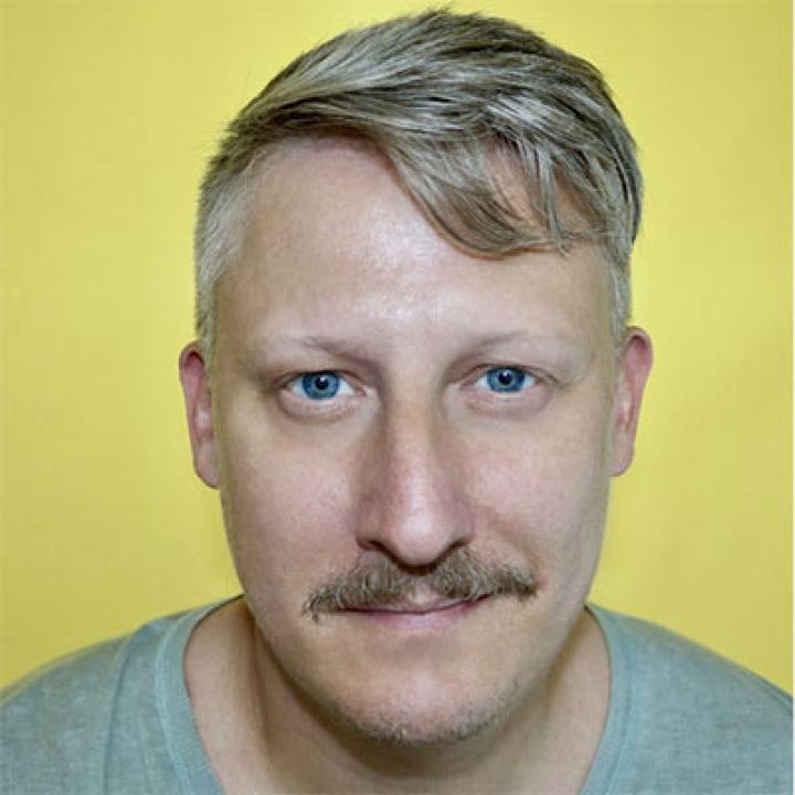 Portrait of Nathan Peck wearing a gray shirt with a yellow background